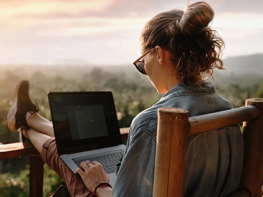 A woman sits relaxed on a balcony, working on a laptop as the sun sets over a lush, green landscape.