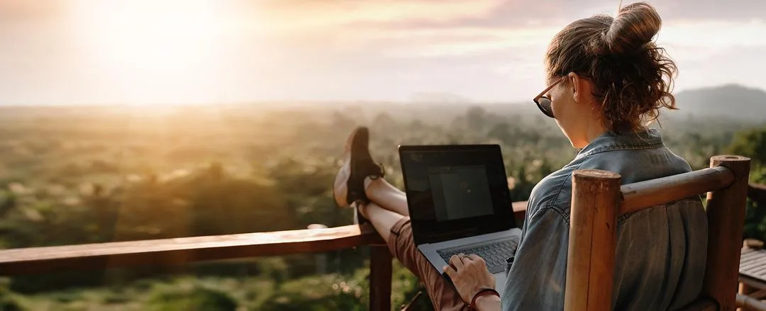 A woman sits relaxed on a balcony, working on a laptop as the sun sets over a lush, green landscape.