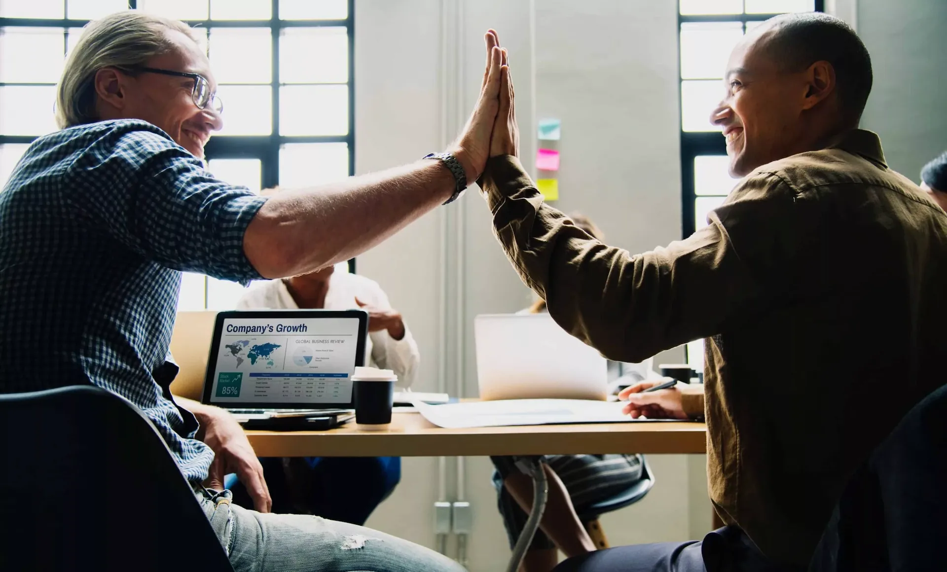Two people at a meeting table share a high-five, celebrating company growth, with a laptop displaying charts in the foreground.