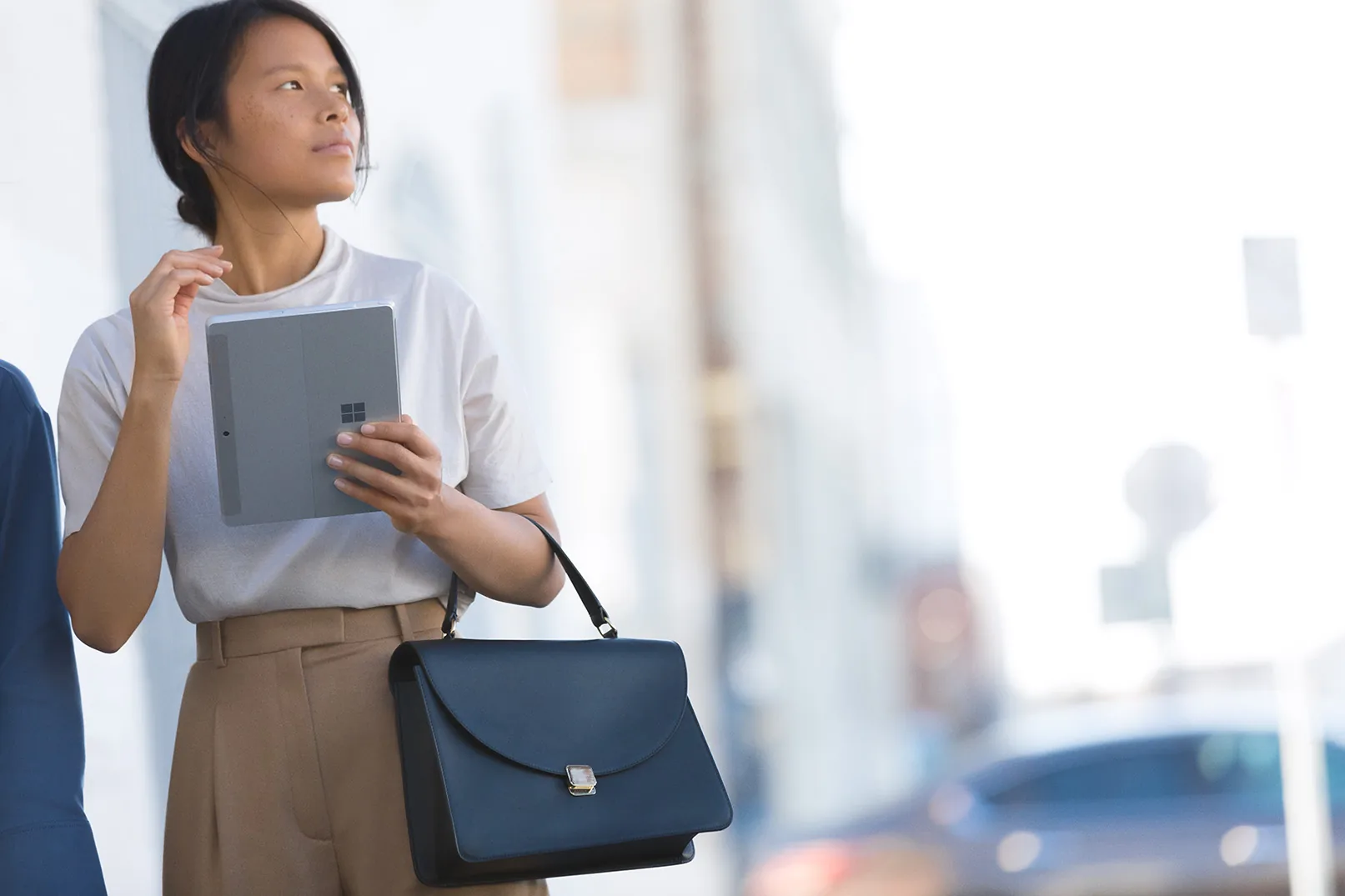 A person stands outdoors, holding a tablet and a stylish black handbag, dressed in a casual white top and beige high-waisted trousers.