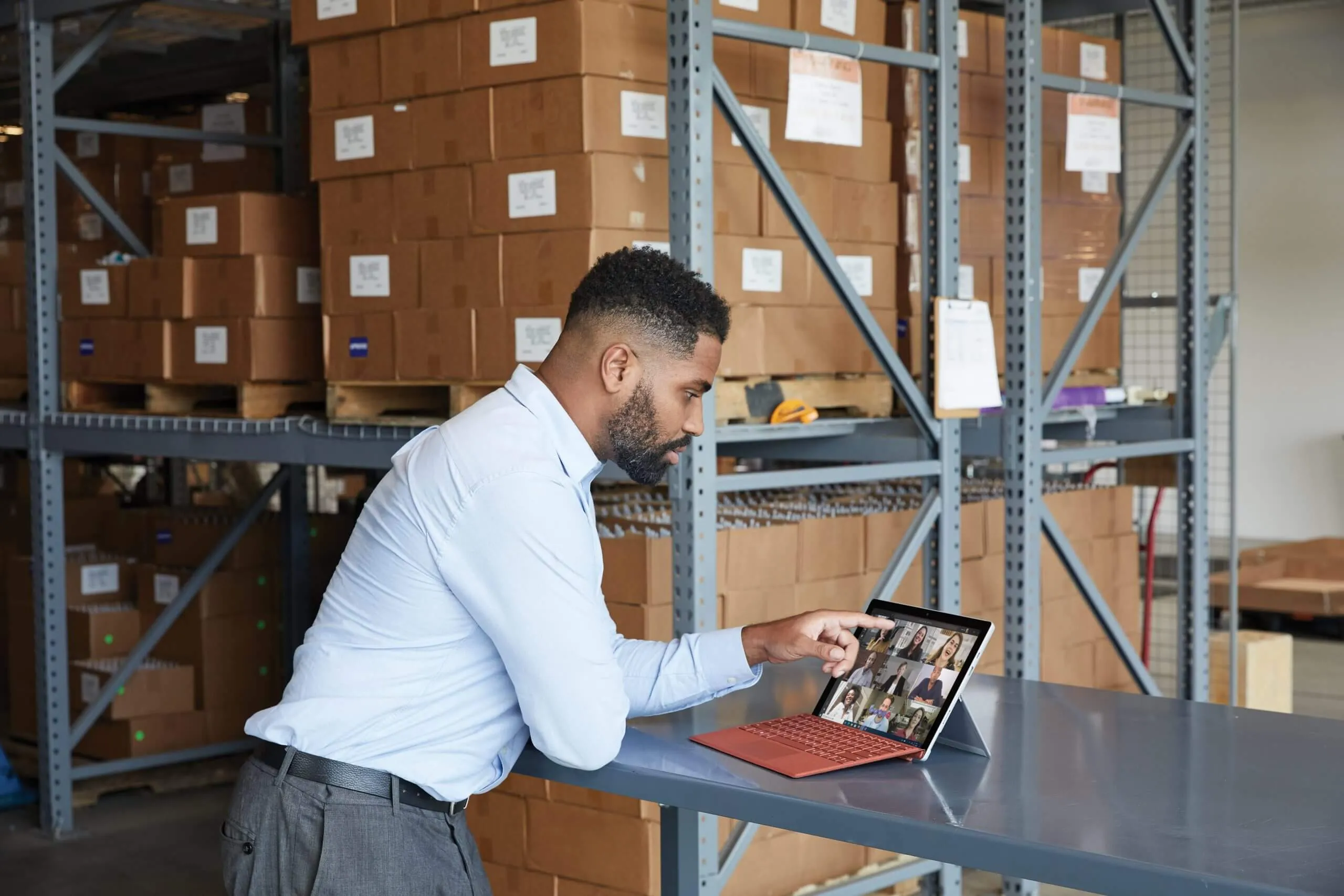 A man in a warehouse leans on a bench, joining a video call on a Surface tablet with a red keyboard, shelves of cartons behind him.