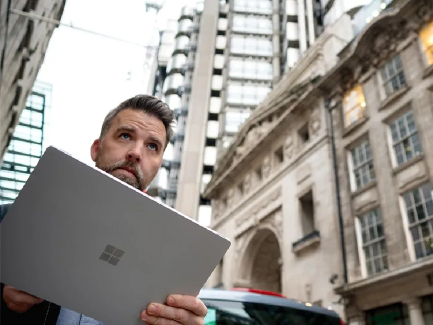 A person holding a Microsoft laptop outdoors, surrounded by tall modern buildings and classic architecture.