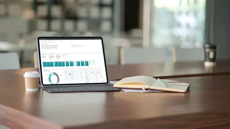 A laptop displaying data analytics on a wooden table, beside a coffee cup and an open notebook.