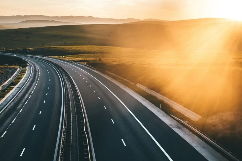 A winding highway stretches through lush green fields under a glowing sunset, with soft light illuminating the landscape.