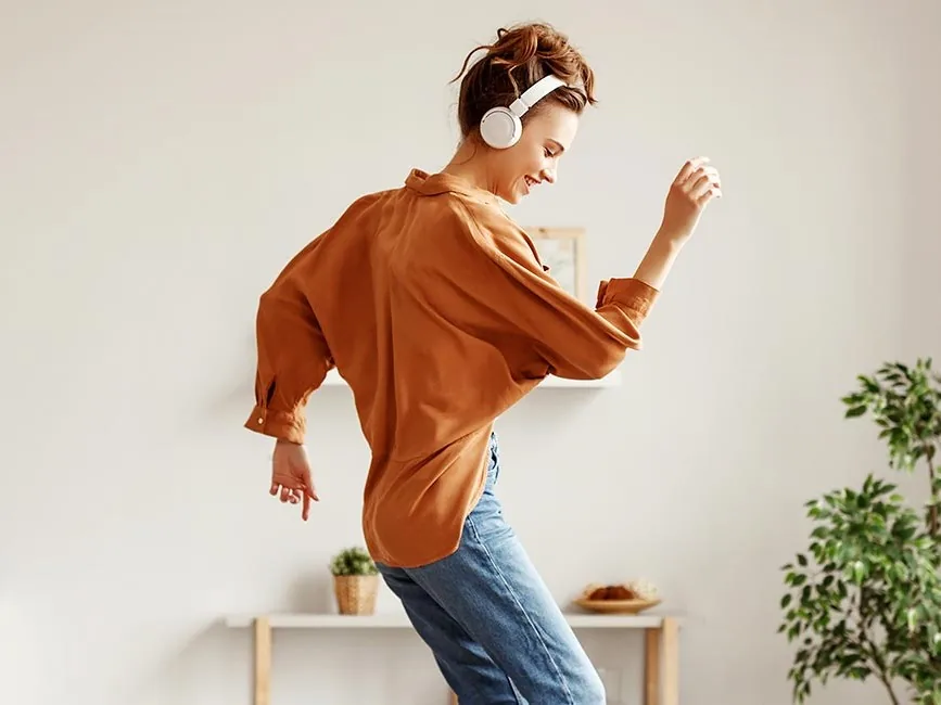 A person in a brown shirt dances joyfully in a cozy, bright room adorned with plants and decorative elements.