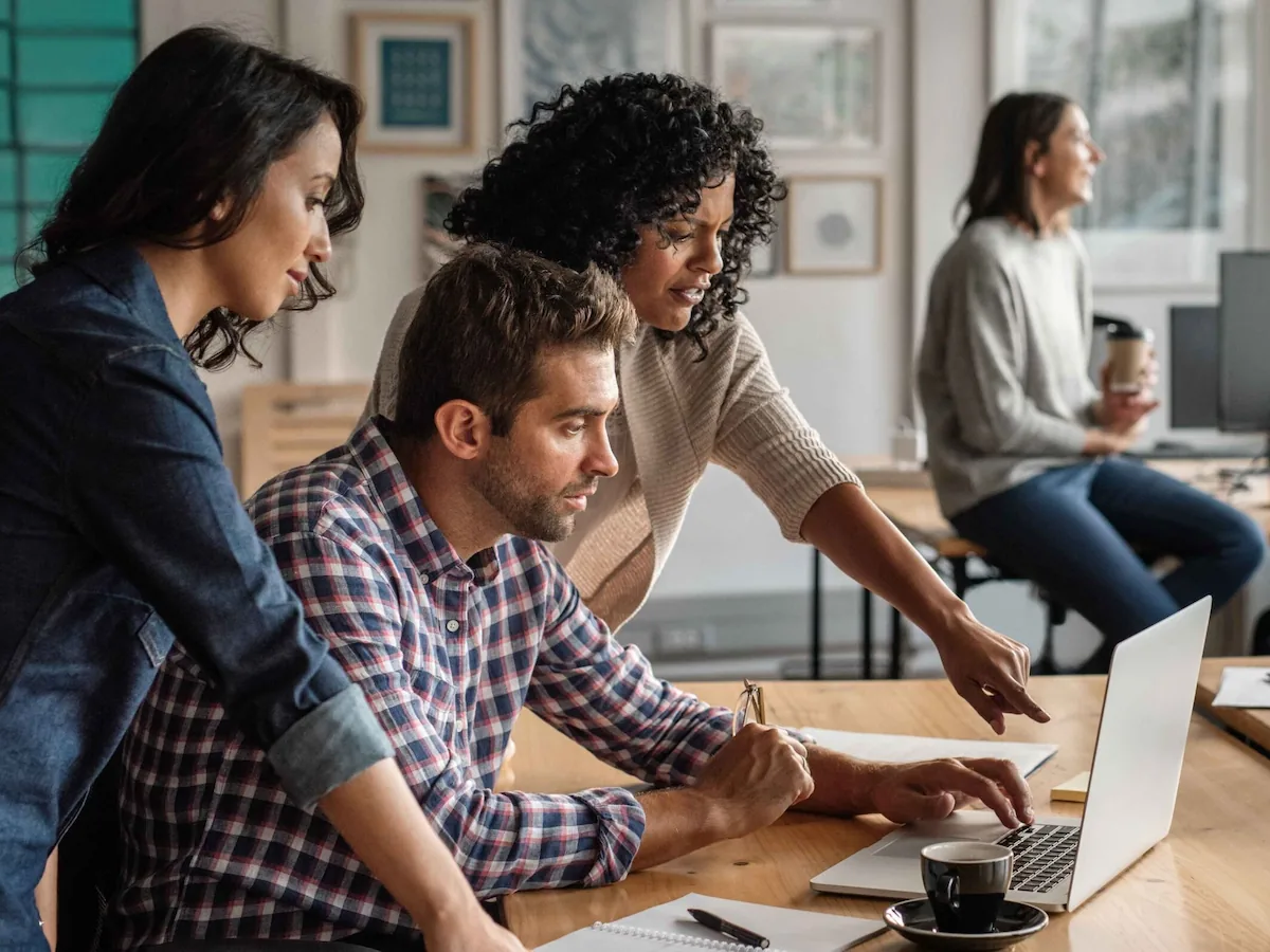 A group of four people collaborates around a laptop on a wooden table, sharing ideas and discussing work in a modern office setting.