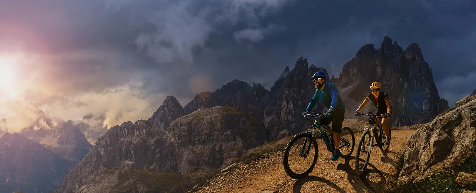 Two mountain bikers ascend a rugged trail surrounded by dramatic mountains under a moody sky, with sunlight breaking through clouds.