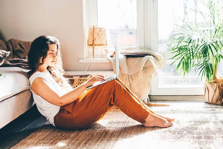 A person sits on a cozy rug, using a laptop in a sunlit living room, surrounded by plants and comfortable furnishings.