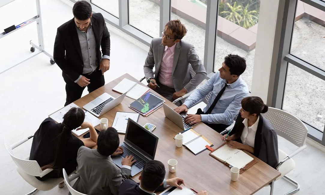 A diverse group of professionals engaged in a meeting around a conference table with laptops and documents.