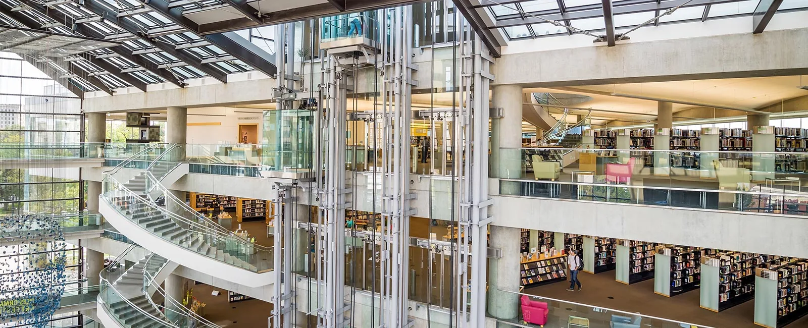 A modern library interior featuring a glass elevator, spiral staircase, and several levels filled with bookshelves.