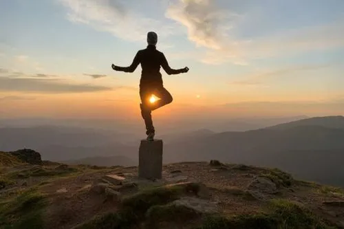 A person silhouetted against a sunrise, balancing on one leg on a stone marker at a mountain summit.
