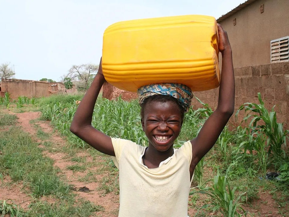 A smiling child balancing a large yellow water container on her head on a dirt path between rows of maize.