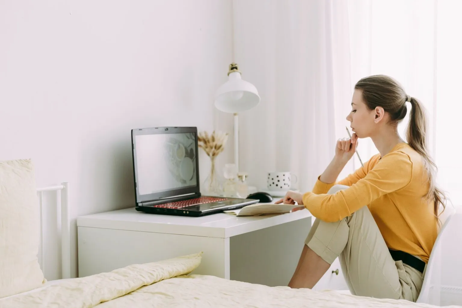 A person in a yellow jumper sitting on a bed, studying a laptop on a small white desk in a sunlit bedroom.