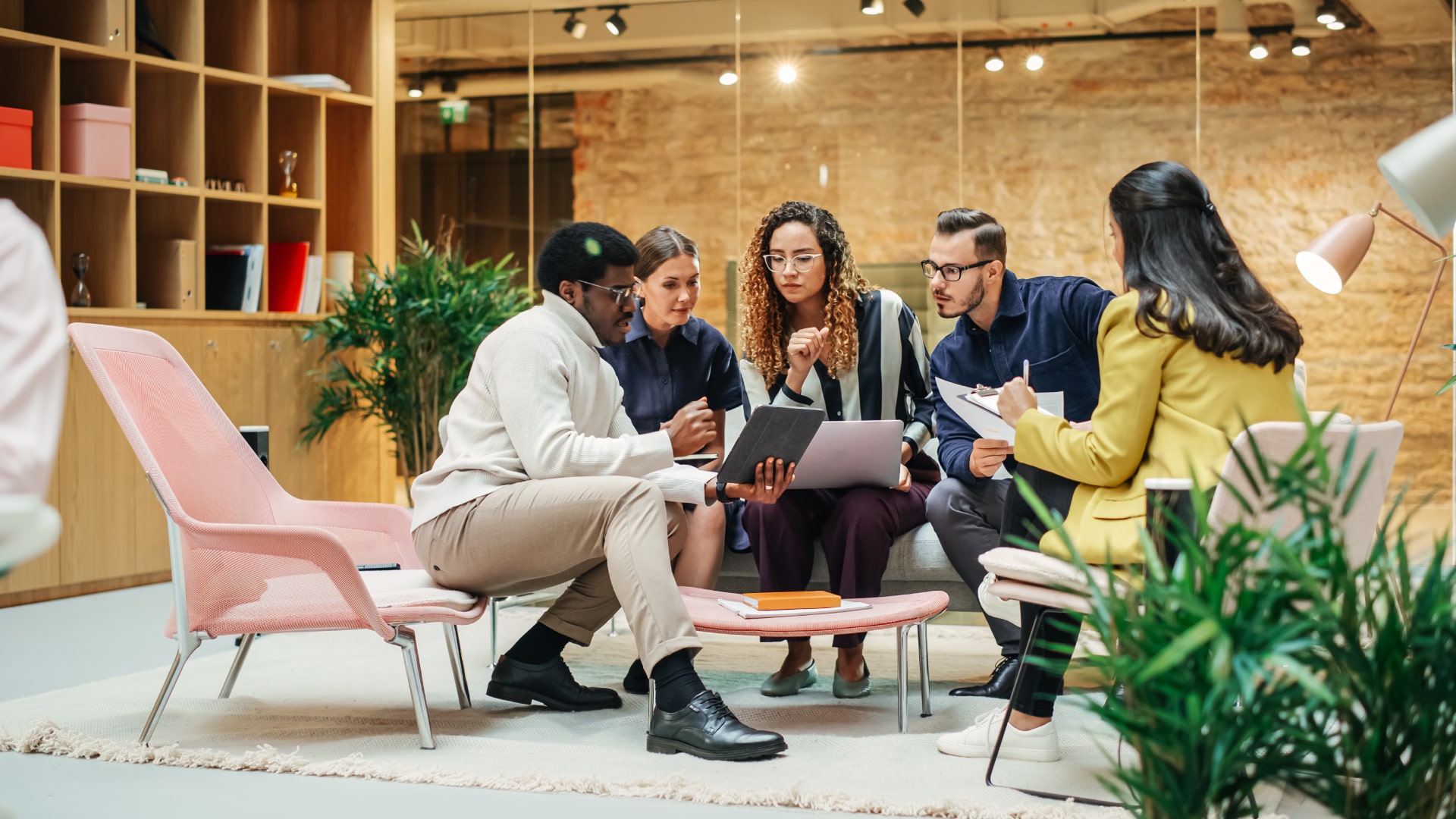 Five colleagues sitting together in a bright, plant-filled office lounge, talking over a tablet and printed notes.